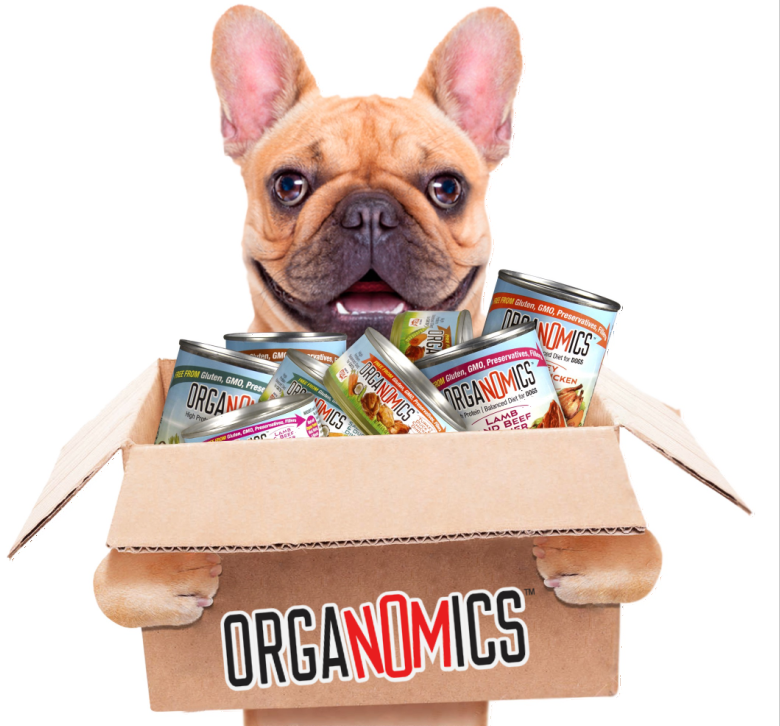 A French Bulldog stands behind a cardboard box filled with OrgaNOMics Salmon and Duck Dinner for Dogs - 12, 12.5 oz cans, with the OrgaNOMics logo printed on the box.