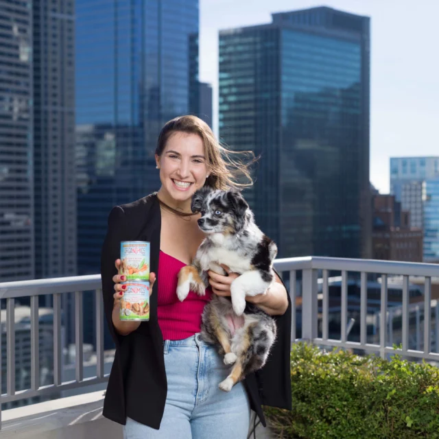 A woman holding a can and a small dog stands on a rooftop with tall city buildings in the background.