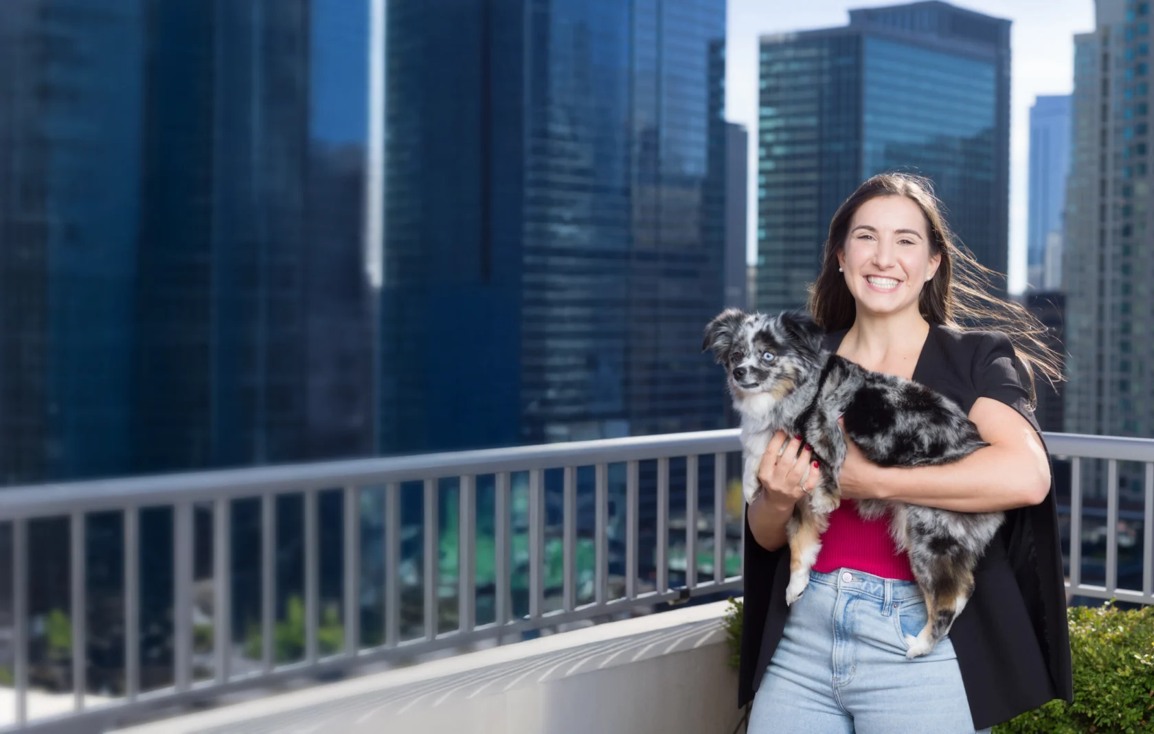 Woman smiling and holding an Australian Shepherd puppy on a balcony with tall city buildings in the background.